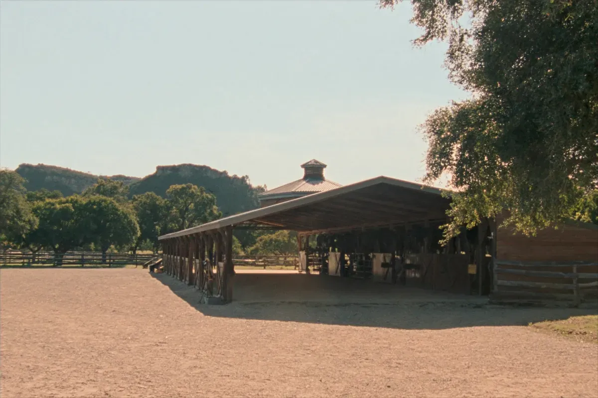 An open shed-row horse stable with a metal roof and limestone bluffs behind in the Texas Hill Country