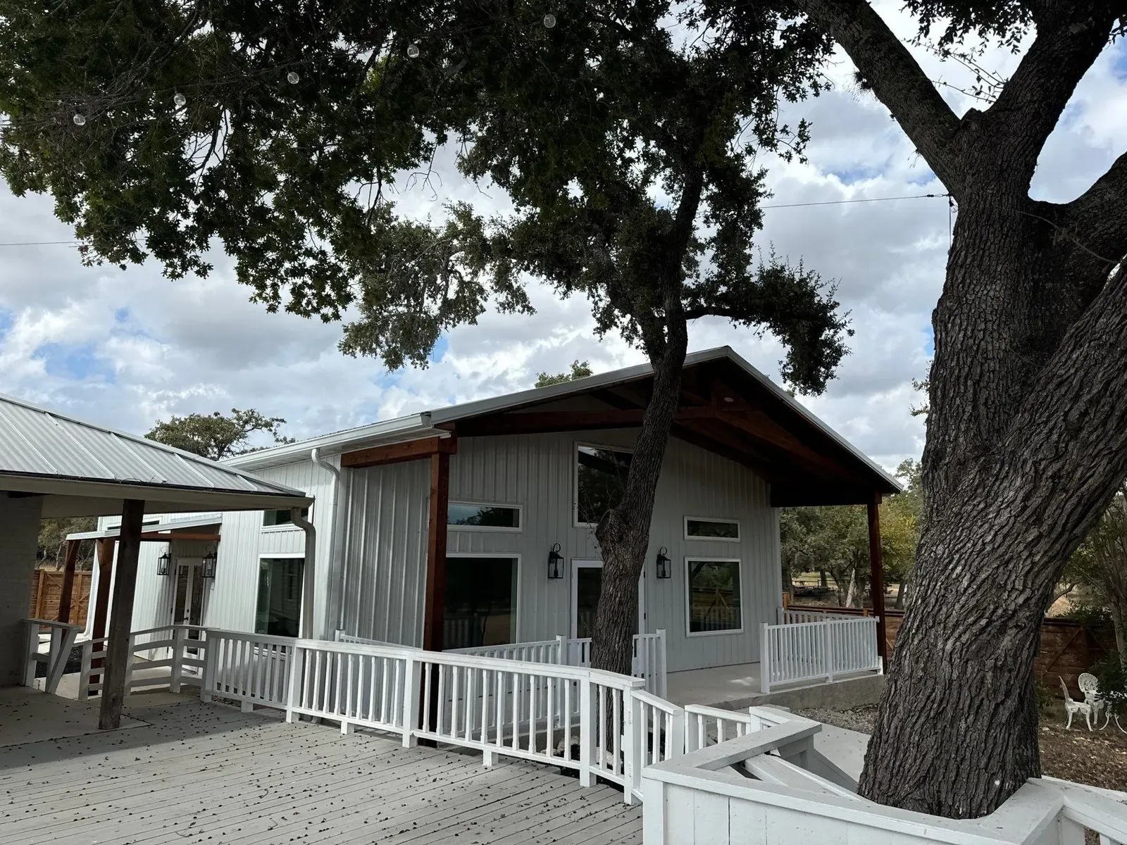 A white board-and-batten barndominium with wood-trimmed gables and a deck built around a large live oak in Texas