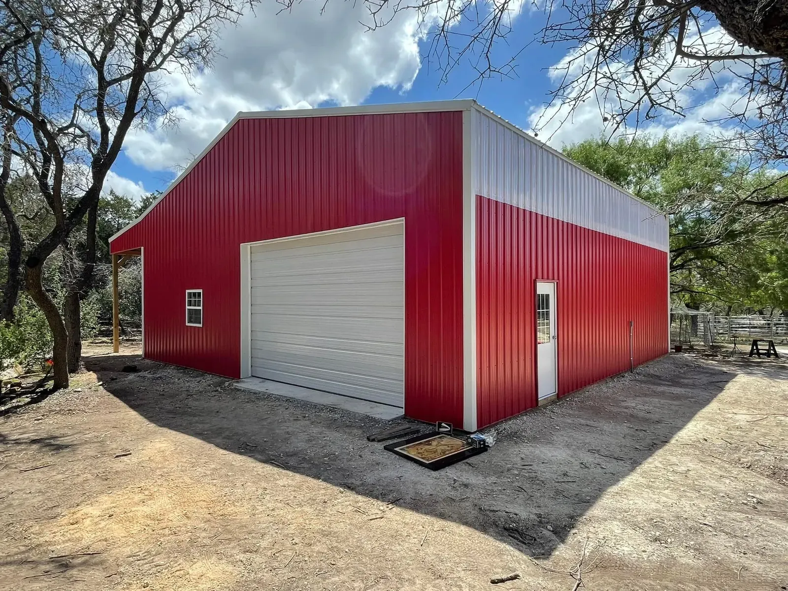 A red and white steel workshop with a roll-up door and a walk door under oak trees in Texas