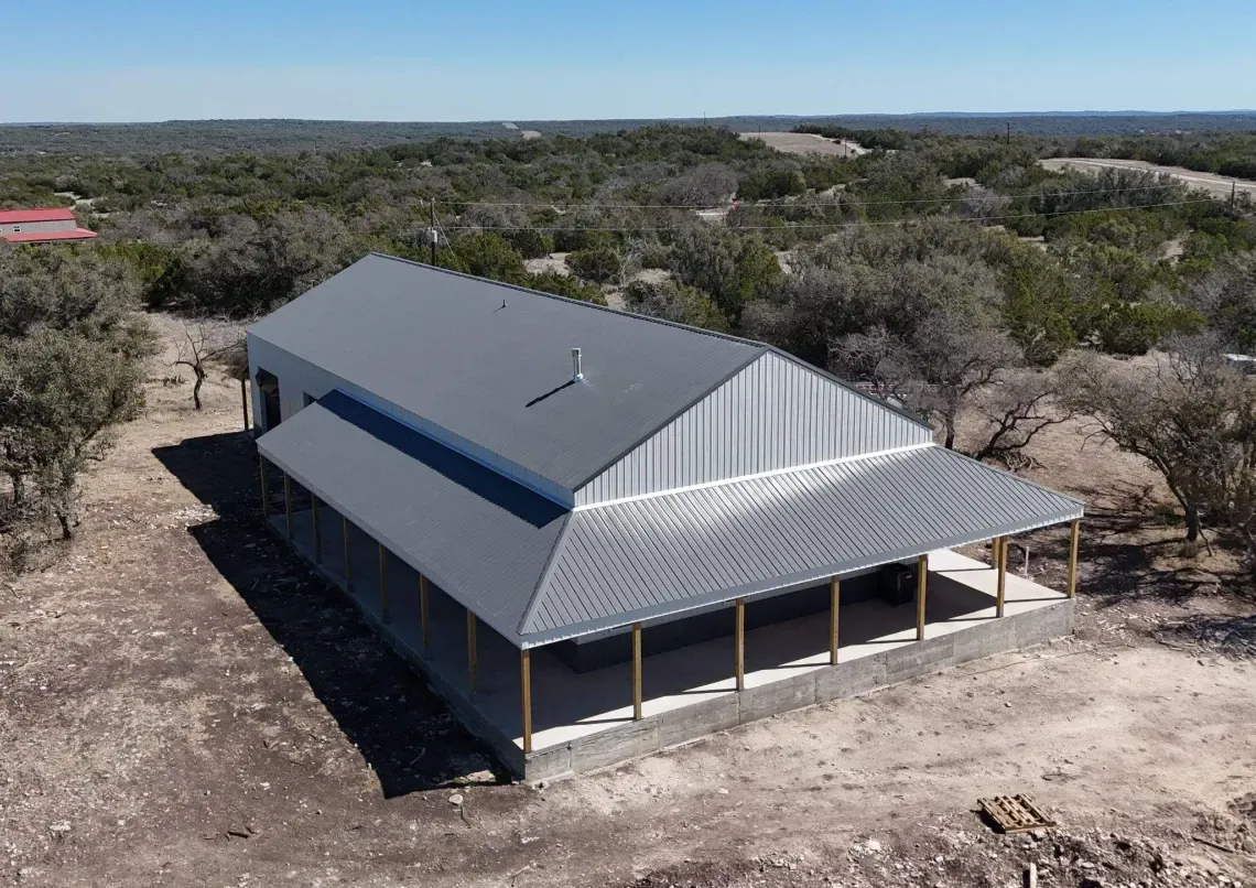 Aerial view of a gray steel building with a wraparound covered porch on a concrete slab among cedar and live oak in Texas